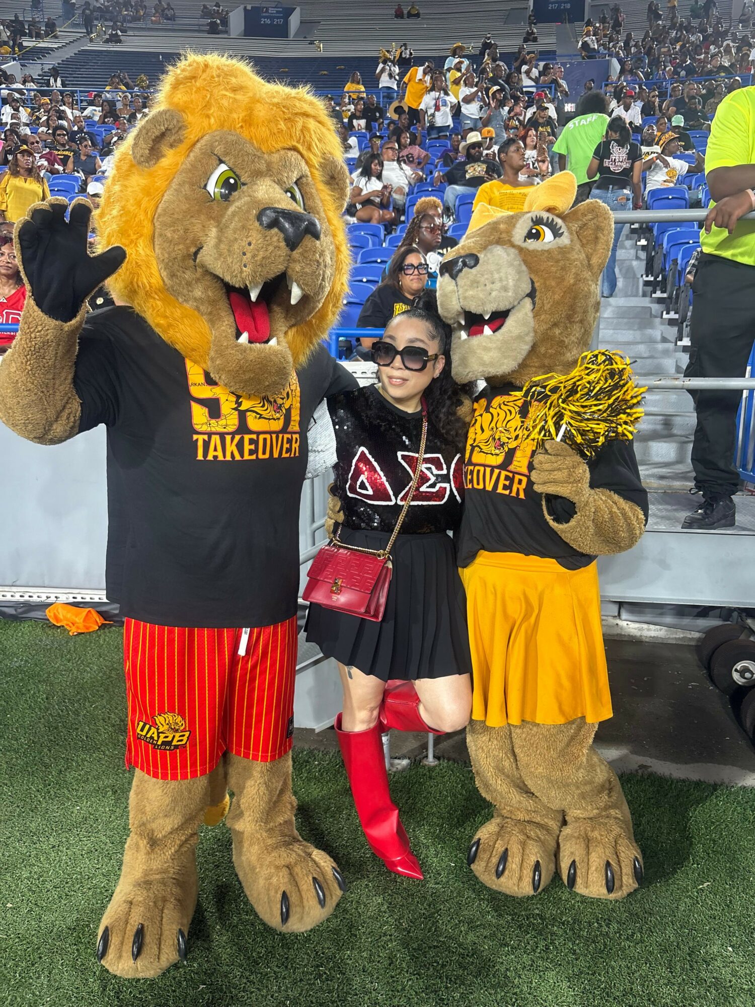 Two people in lion mascot costumes pose with a woman at a sports event, one mascot wears a black shirt, the other a yellow shirt.