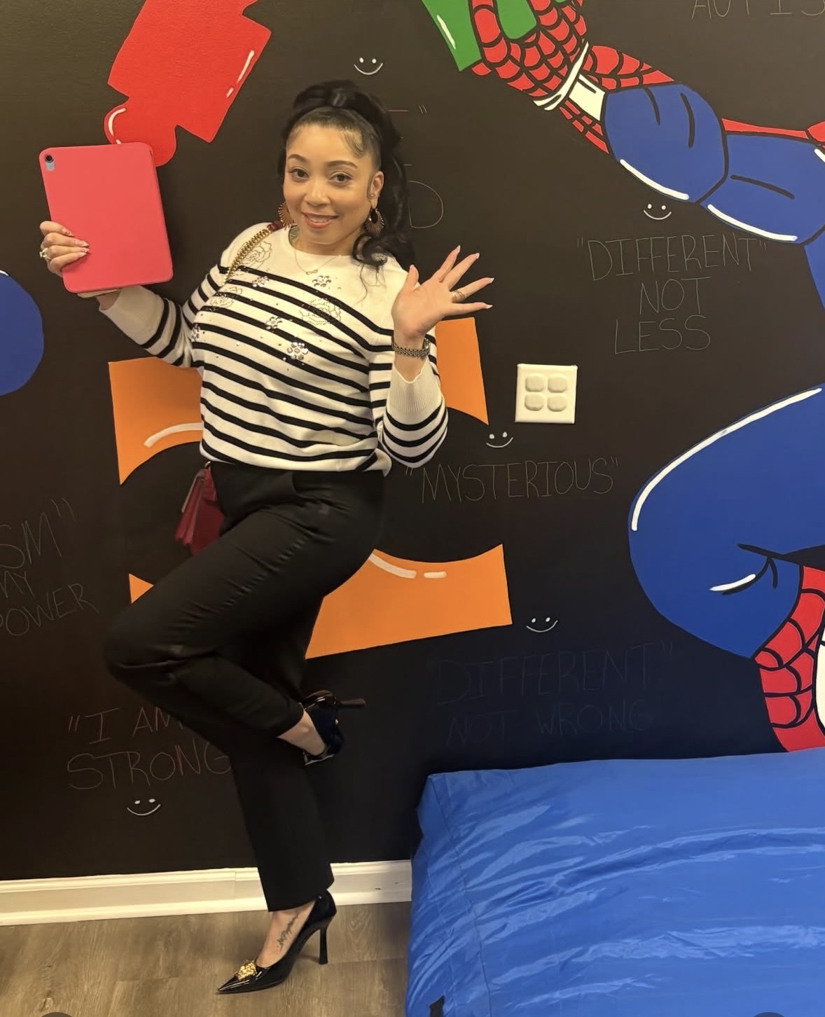 Woman sitting on a bed, waving, holding a pink tablet, in a room with a superhero-themed wall mural.
