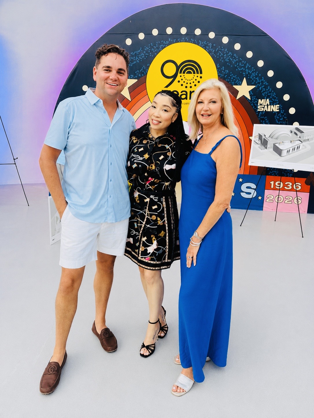 Three people standing together in front of a circular backdrop with a yellow logo, smiling, with a table and pink chair nearby.
