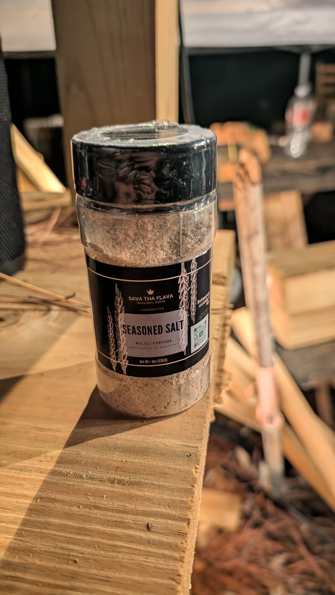 A black-labeled jar of seasoned salt sits on a wooden surface in a warmly lit room.