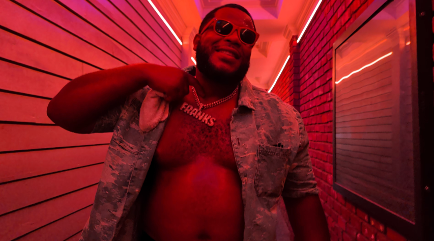 Person wearing sunglasses and an open shirt stands in a narrow, red-lit corridor.