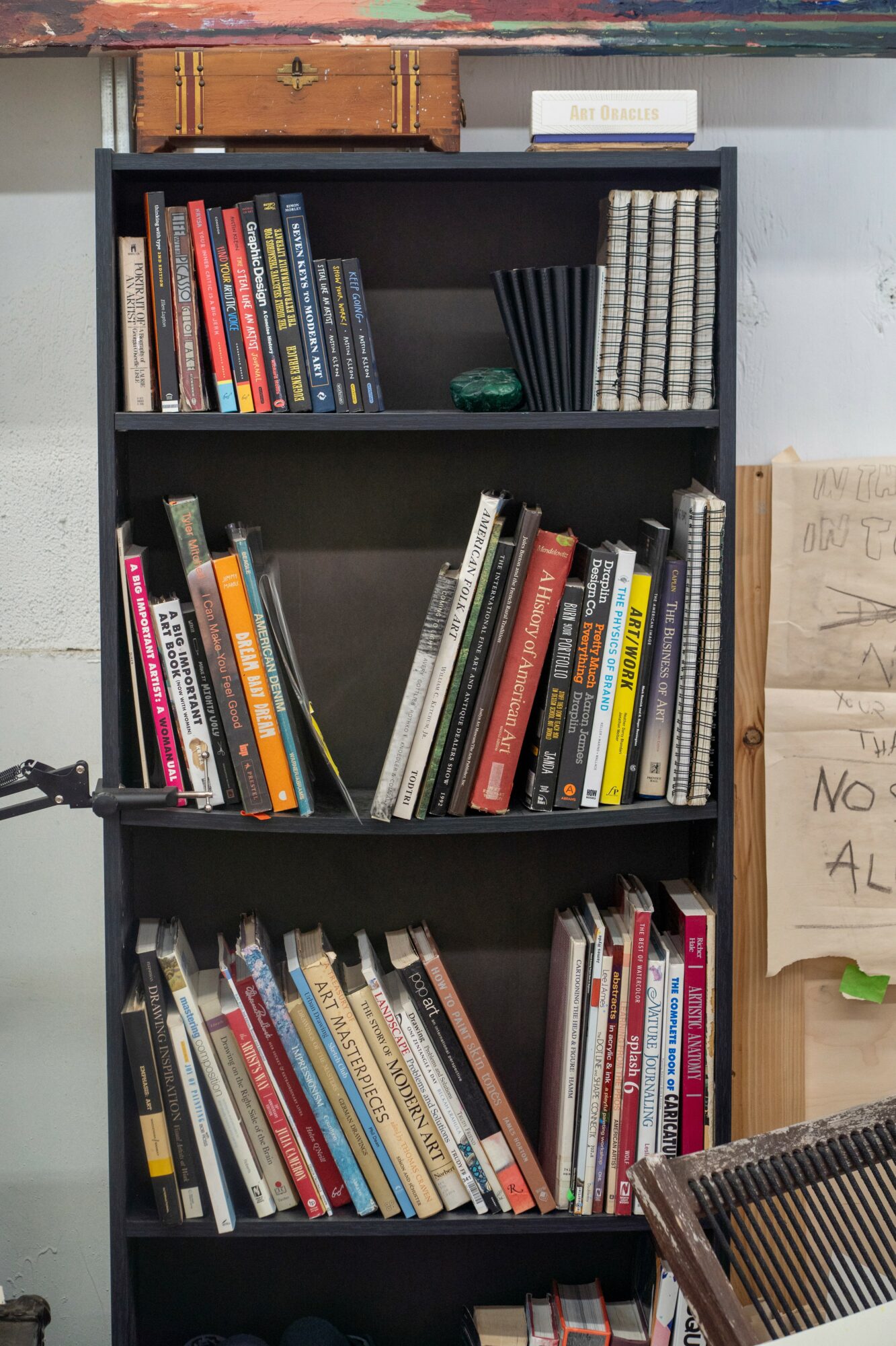 Black bookshelf filled with books, some stacked vertically and others leaning, against a white wall.