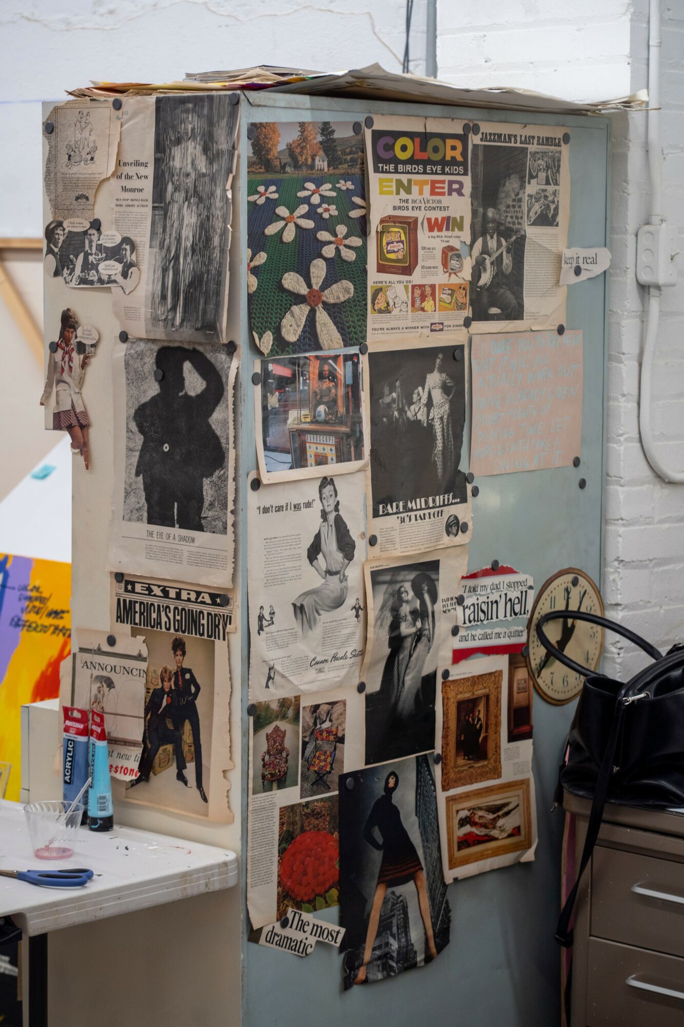 Wall covered with posters, photographs, and artwork, with a desk and a black bag nearby.