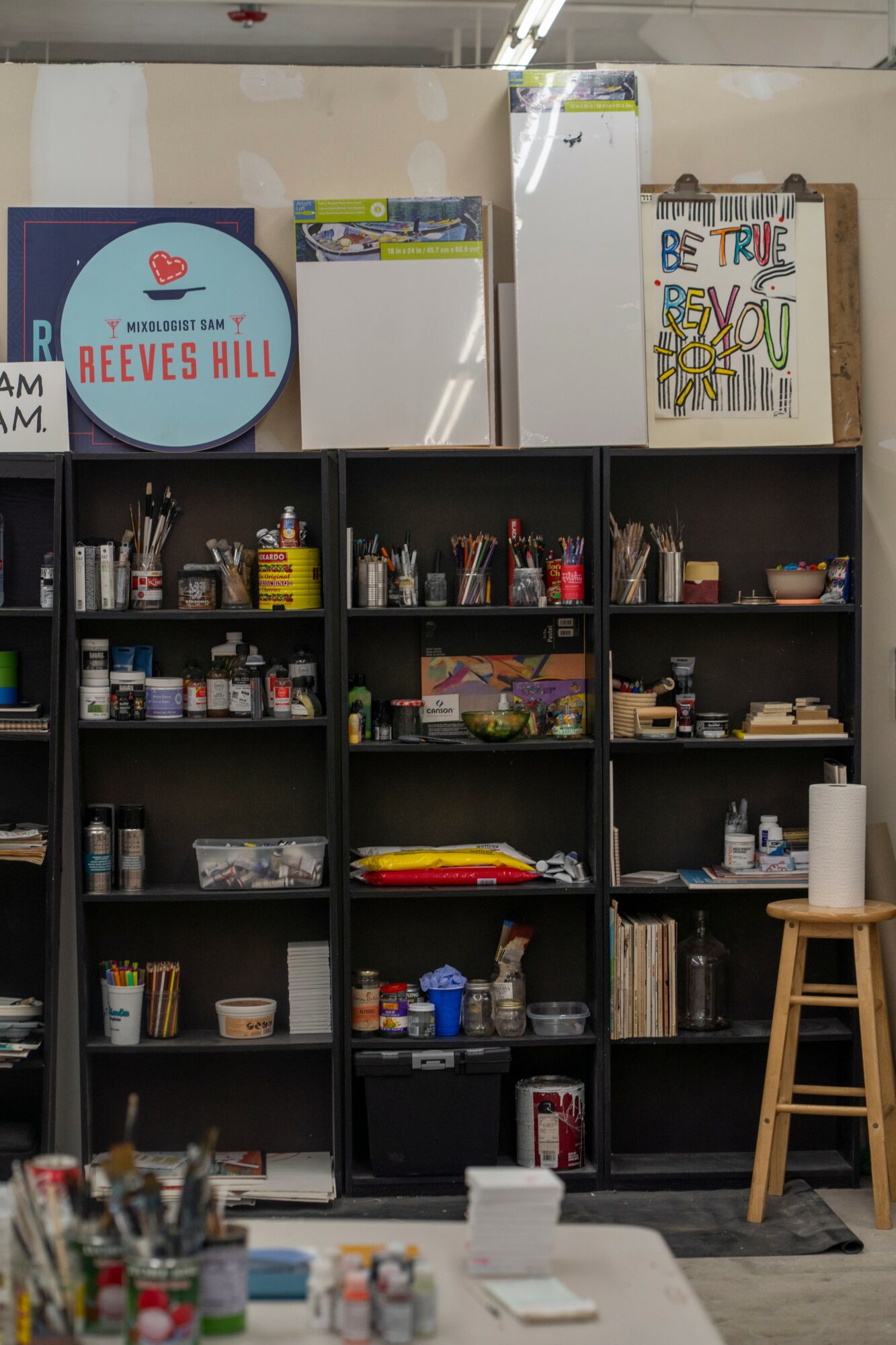 Black shelving unit filled with art supplies and books, with a wooden stool beside it, in a classroom or studio.