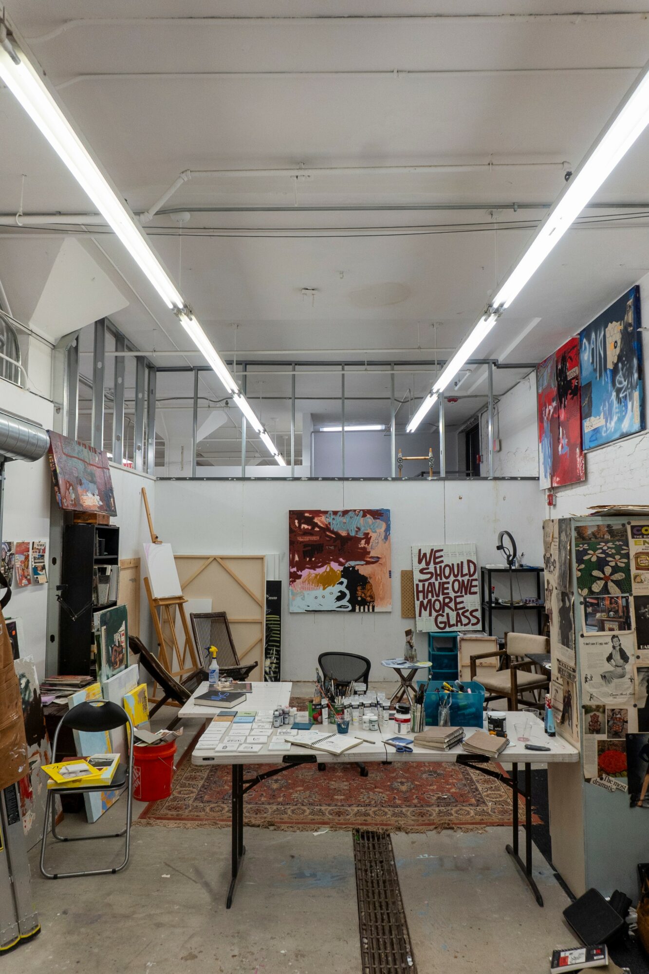 Art studio with tables, chairs, paintings, and art supplies, illuminated by overhead fluorescent lights.