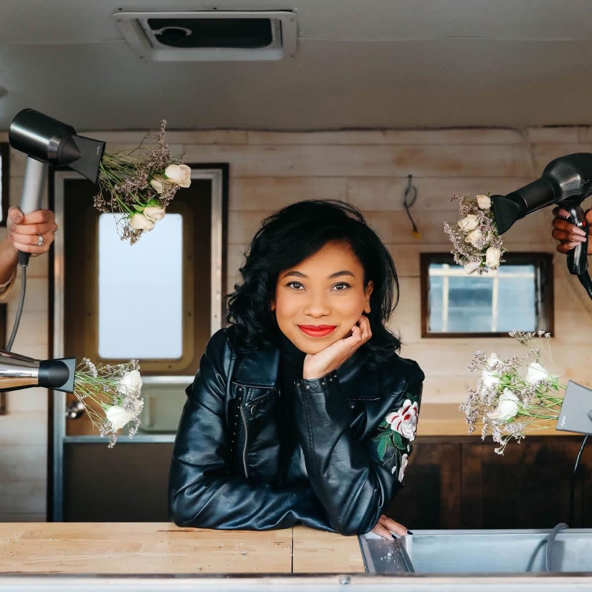 Woman with curly hair and red lipstick sitting at a table, surrounded by four hands holding hairdryers with floral attachments, smiling.