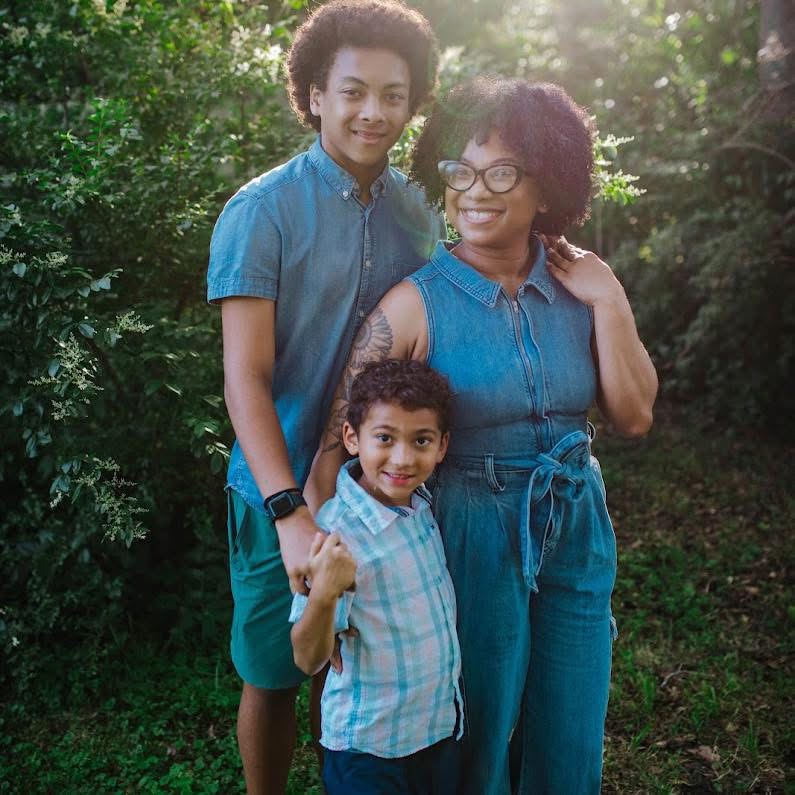Three people, two children and one adult, standing outdoors in a green, wooded area, smiling at the camera.