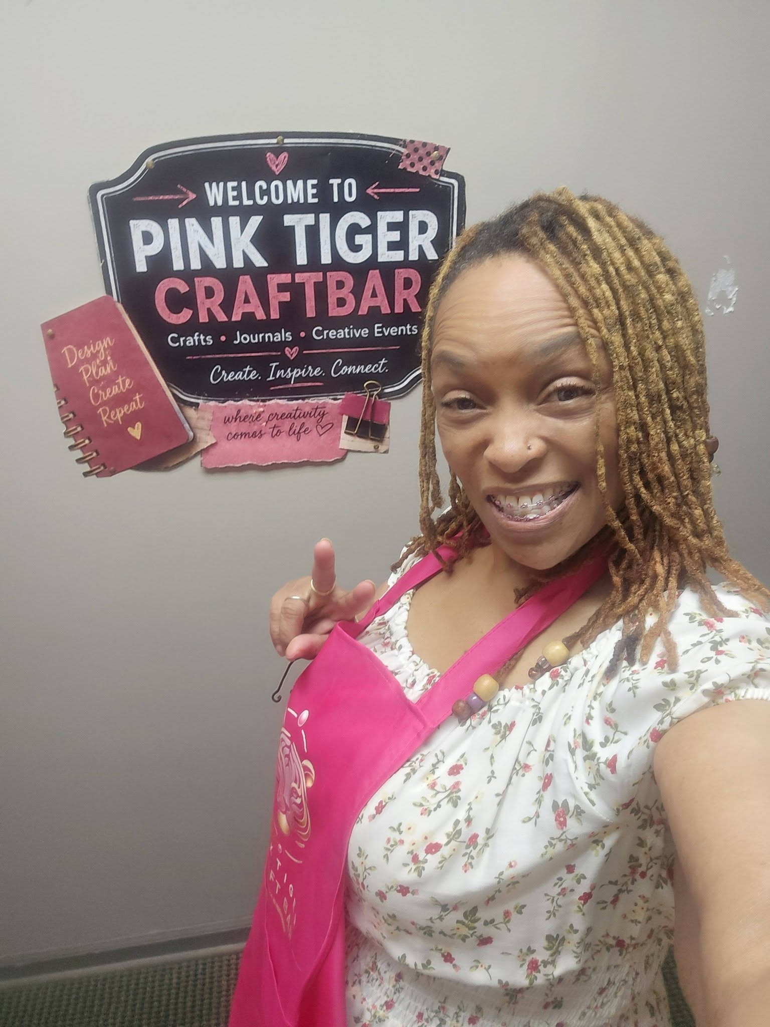 Woman smiling in front of a sign that reads 'Welcome to Pink Tiger Craftbar,' wearing a pink apron and floral top.