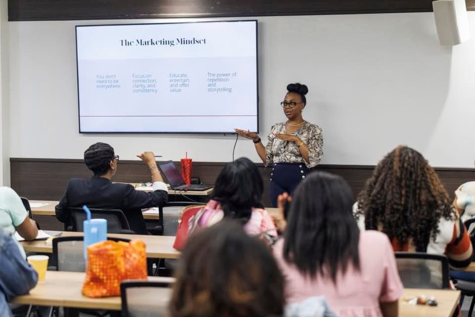A woman presents in front of a classroom with students seated at desks, a large screen behind her displays a presentation slide.