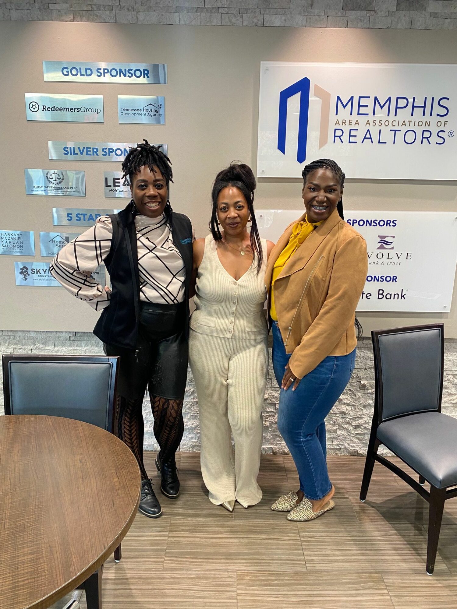 Three women standing in an office, two smiling, one with a serious expression, behind a table and chairs.