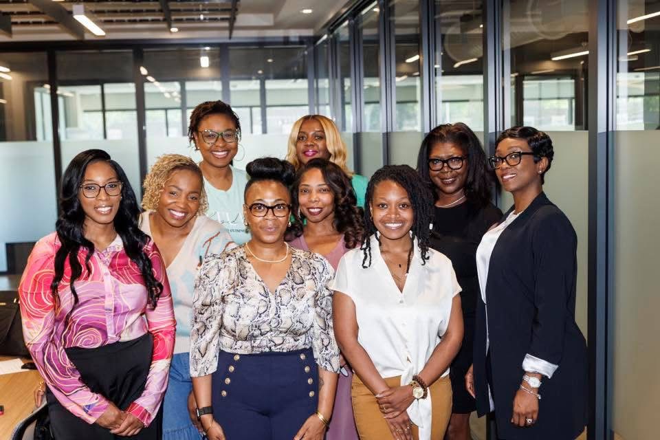 Group of nine women smiling in an office setting with glass walls and modern decor.