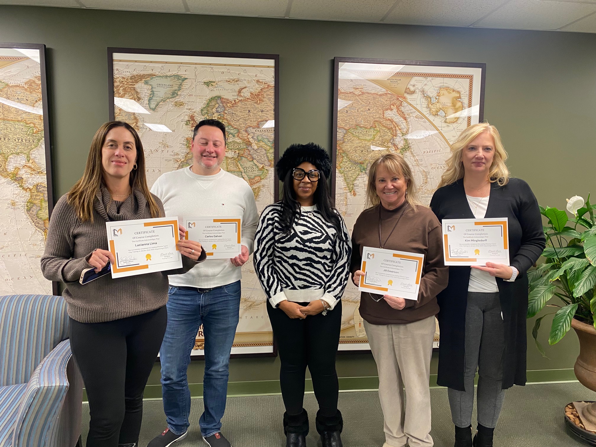 Five people standing indoors holding certificates, with world maps on the wall behind them.