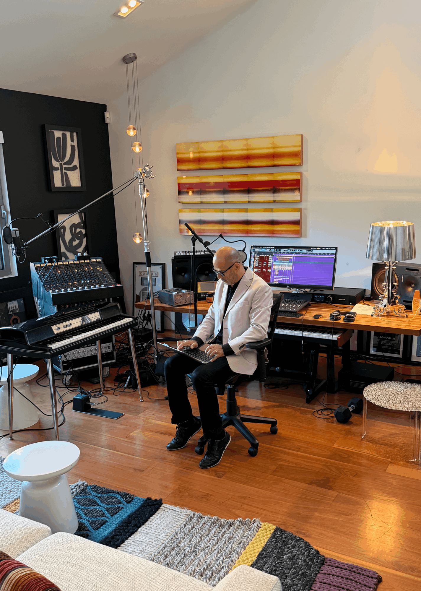 Man in white jacket sitting on chair in a music studio with equipment and colorful wall art behind him.