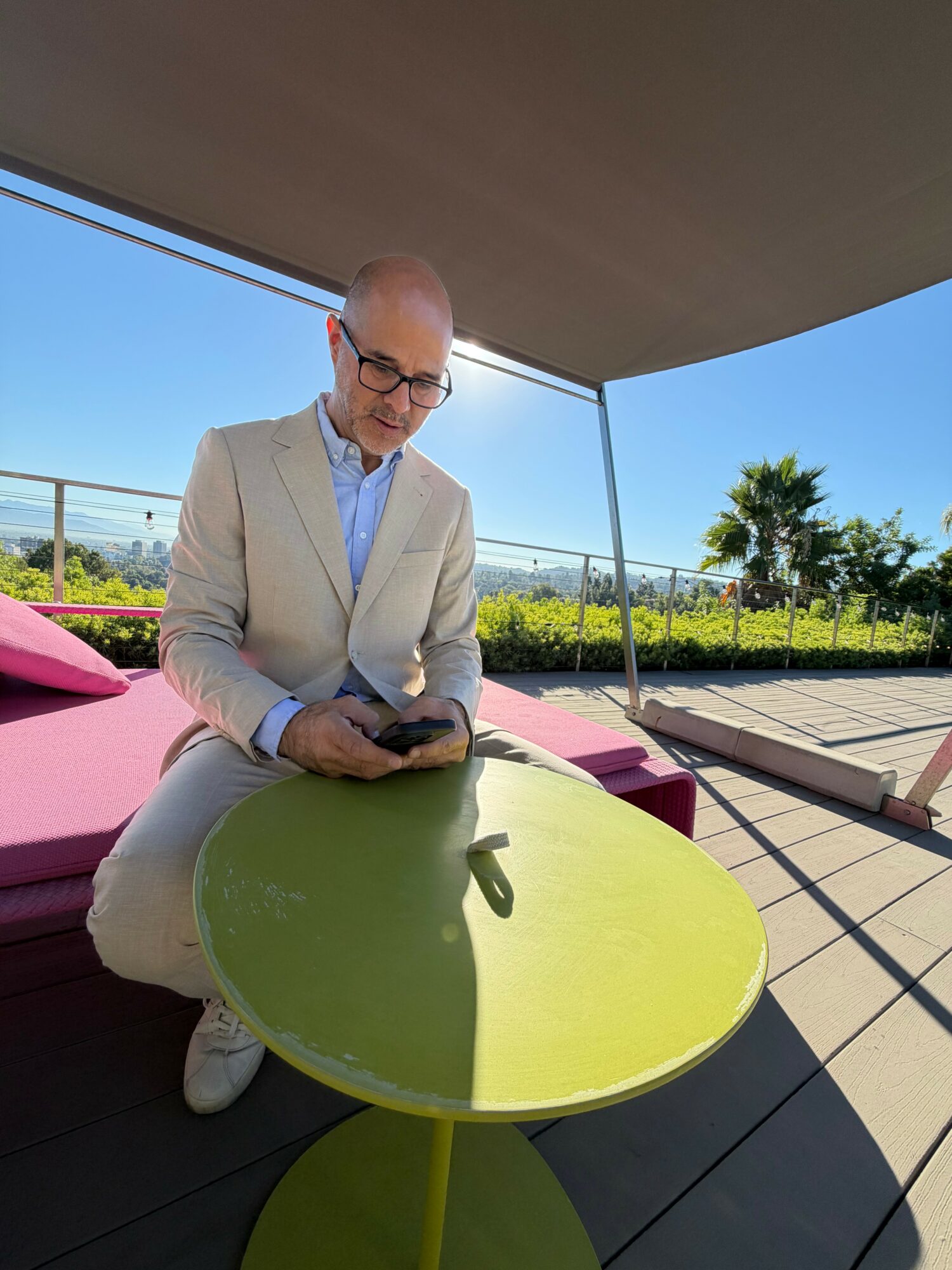 Man in beige suit using a smartphone outdoors on a sunny day, with a green table and pink seating nearby.