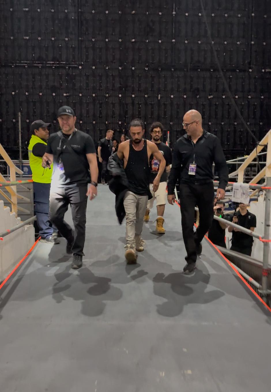 Three men walking on a bridge indoors, with a large black structure in the background.