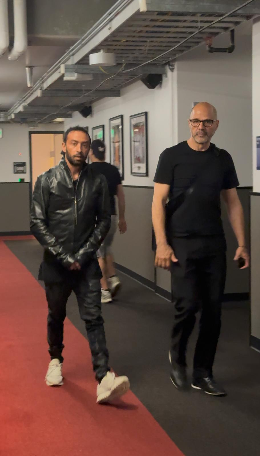 Two men walking in an indoor corridor with framed pictures on the wall, one wearing a leather jacket and the other in black shirt.