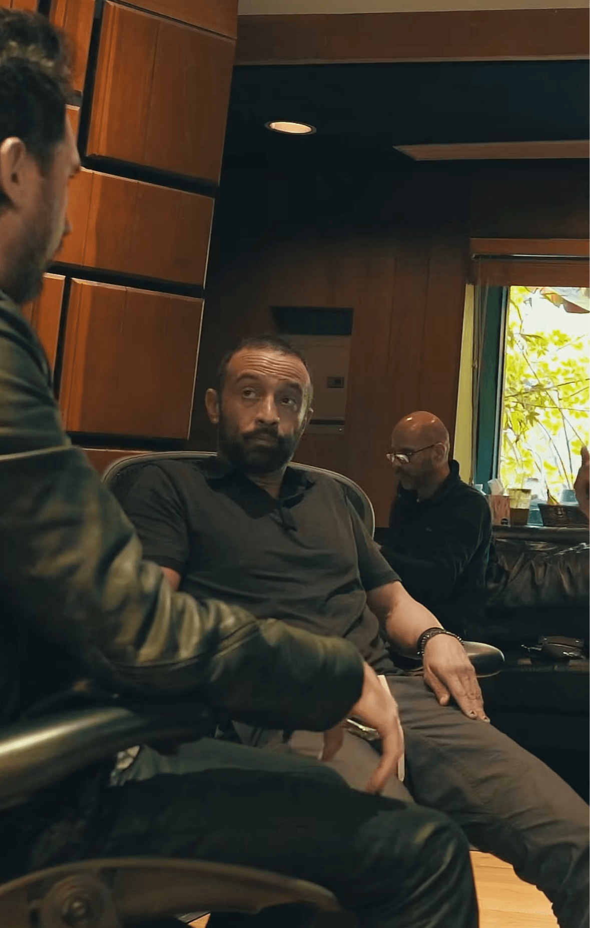Three men sitting in a room with wooden walls and a window showing trees outside.