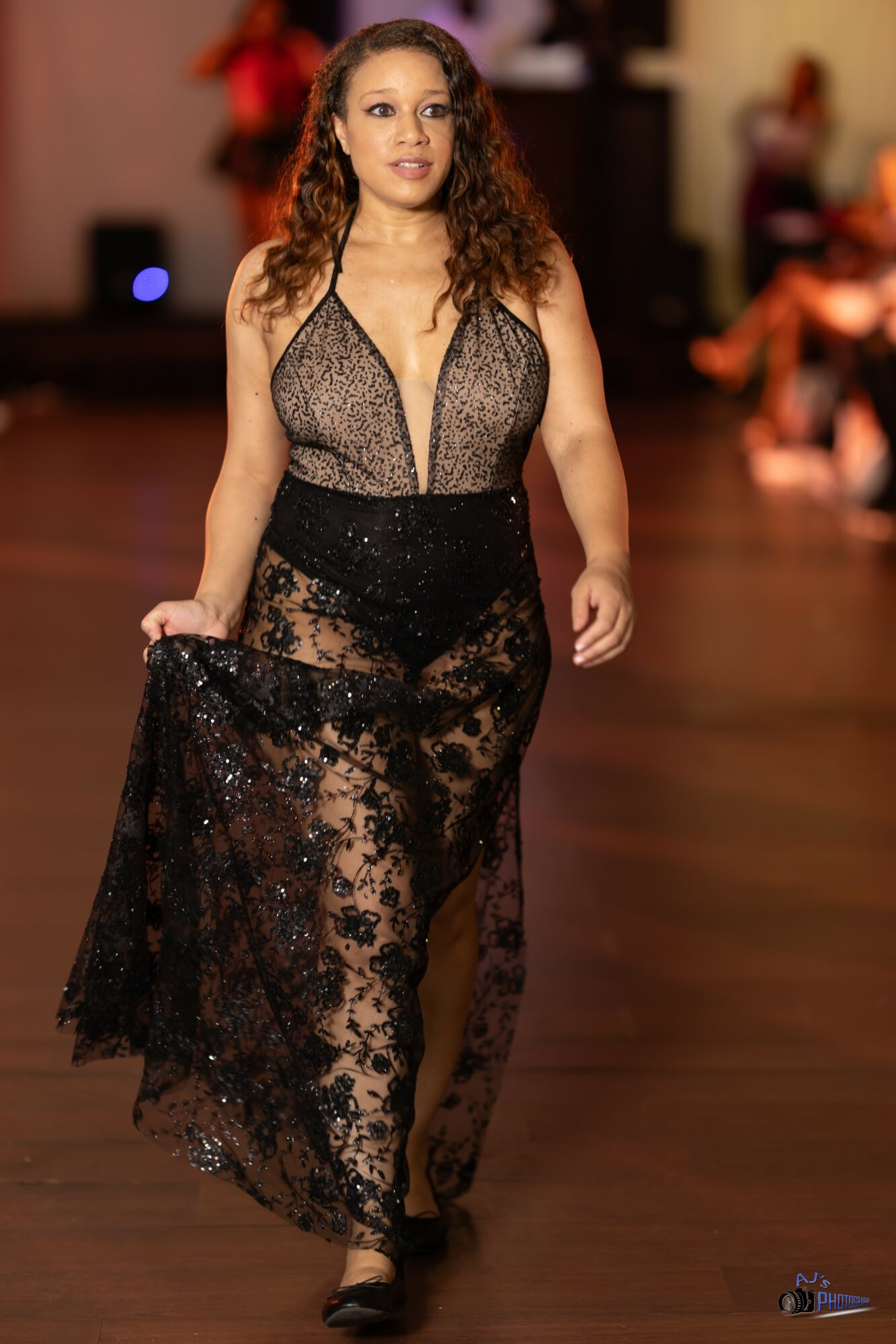 Woman walking on a wooden floor in a black lace dress with a sheer top and long skirt, in an indoor setting.