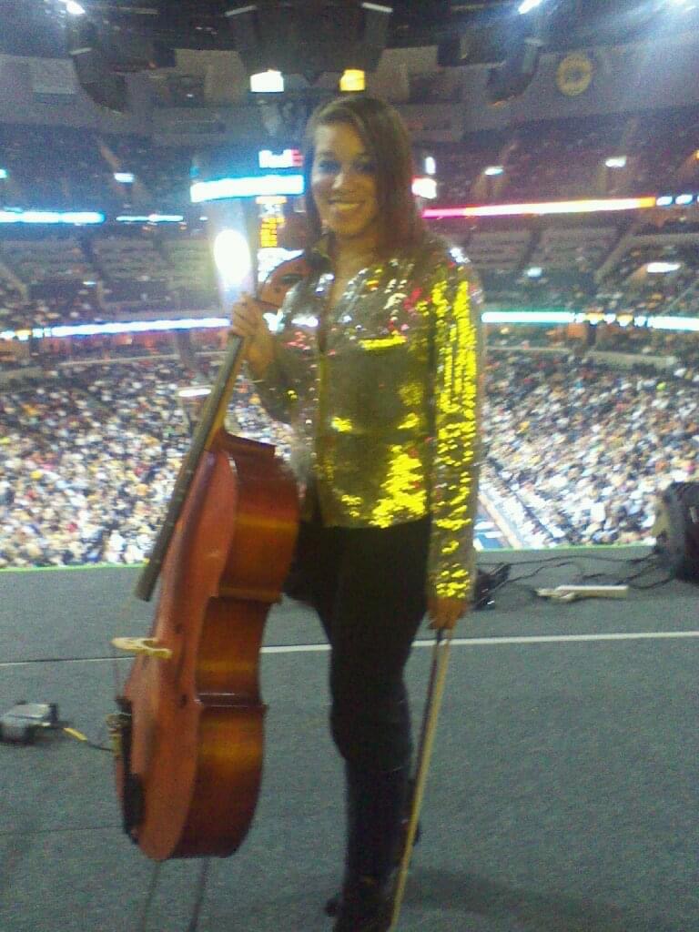 Woman standing on stage with a cello, in a large indoor arena with a crowd, wearing a shiny gold jacket.