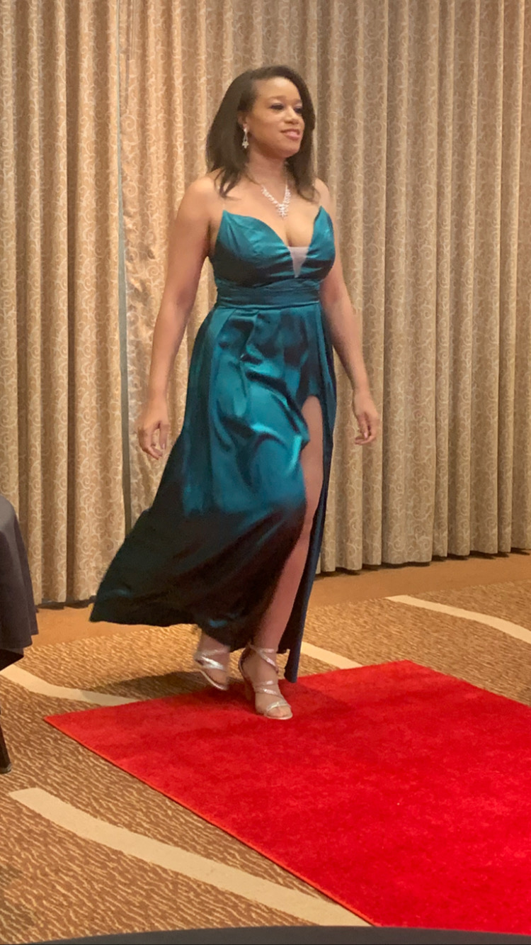 Woman in teal gown walking on red carpet with gold curtain background, wearing high heels and jewelry.