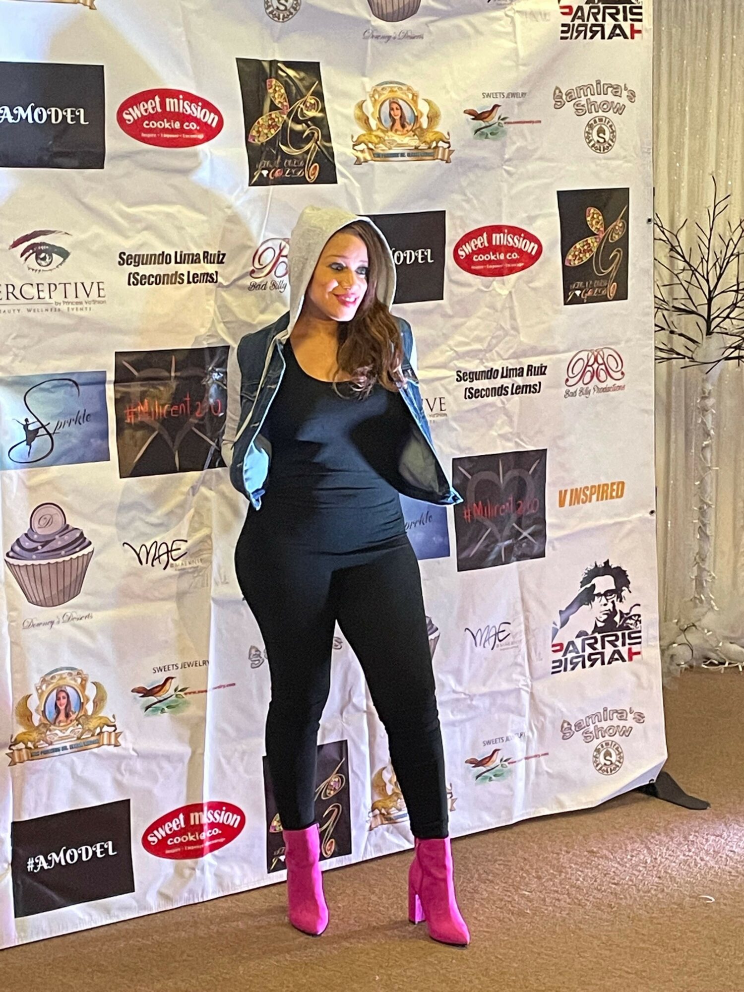 Woman in black outfit and pink shoes standing in front of a promotional backdrop, smiling, with arms raised.