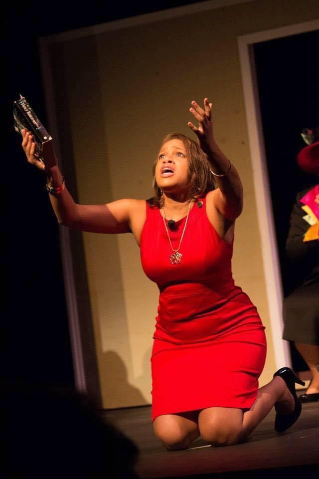 Woman in red dress kneeling on stage, holding a gun in each hand, looking upward with an expressive face.