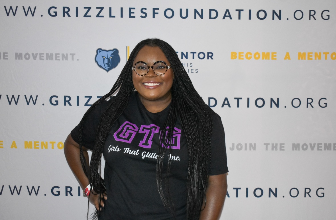 Smiling woman with long braids and glasses standing in front of a backdrop with text and logos.