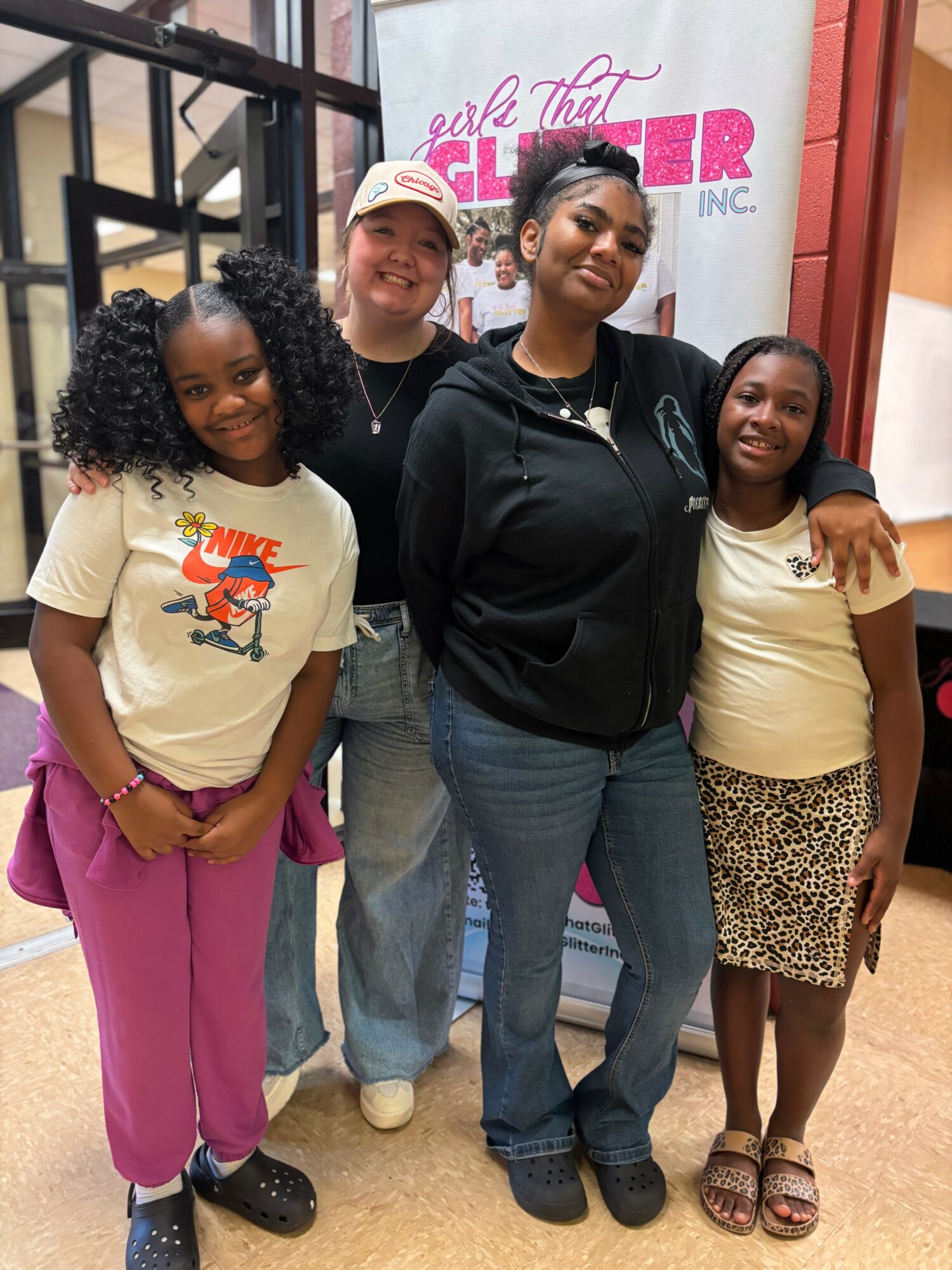 Four women standing together indoors, smiling, with a pink and white sign in the background, wearing casual clothing.