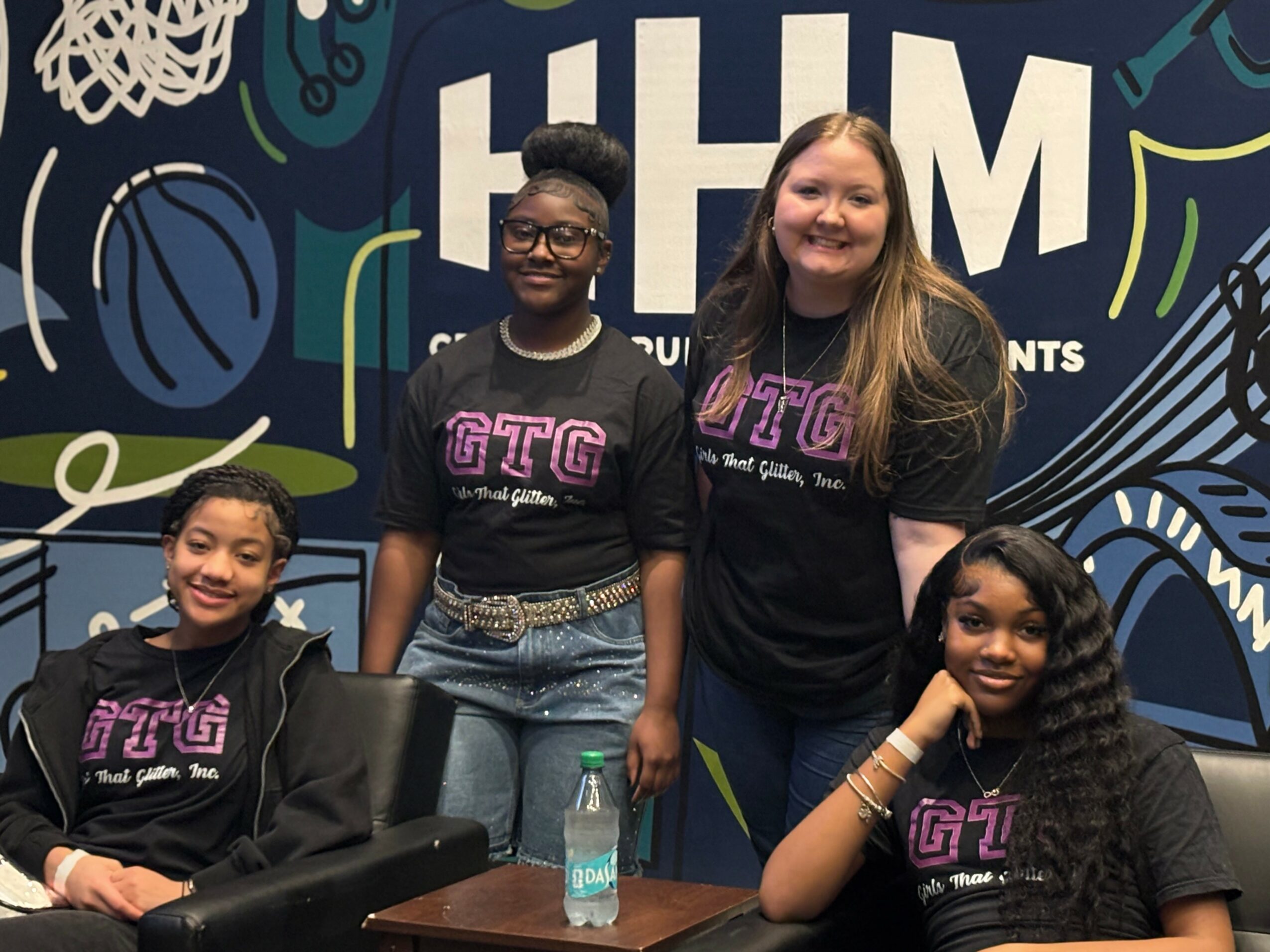 Four smiling young girls wearing matching black T-shirts with pink and white text, standing and sitting in front of a sports-themed backdrop.