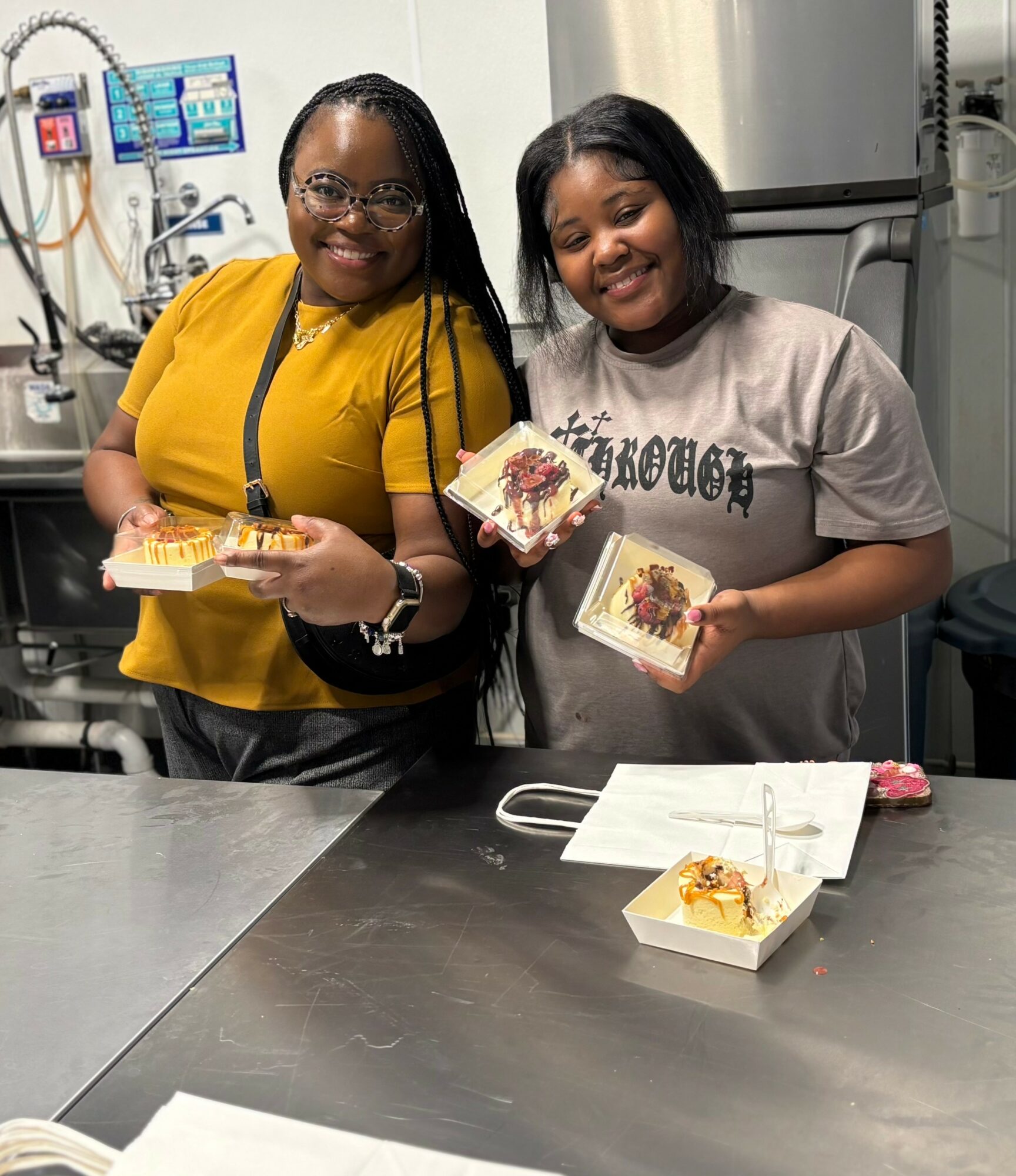 Two smiling women holding food containers in a kitchen, with a box of food on the counter in front of them.