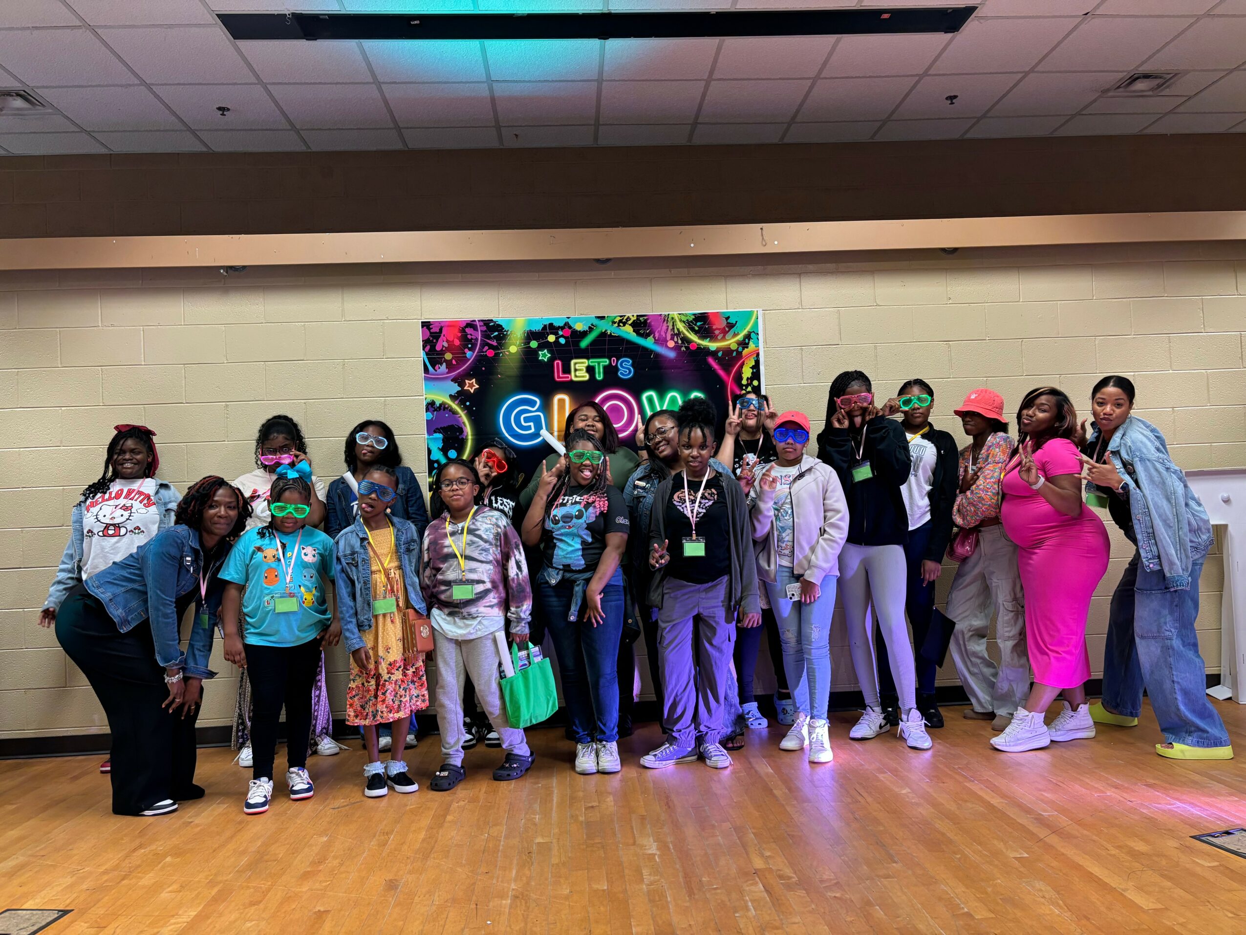 Group of children and adults standing in front of a colorful sign, indoors with a wooden floor and ceiling tiles.