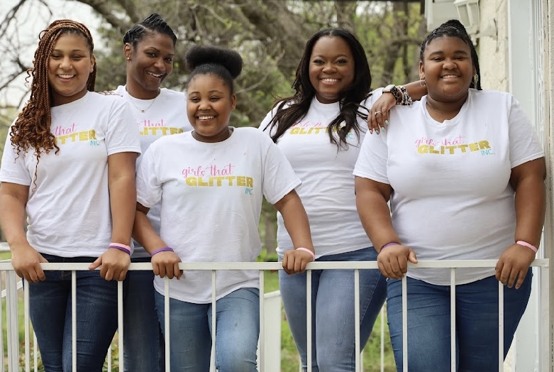 Five women stand outdoors behind a white railing, smiling, wearing white T-shirts and jeans, with trees in the background.