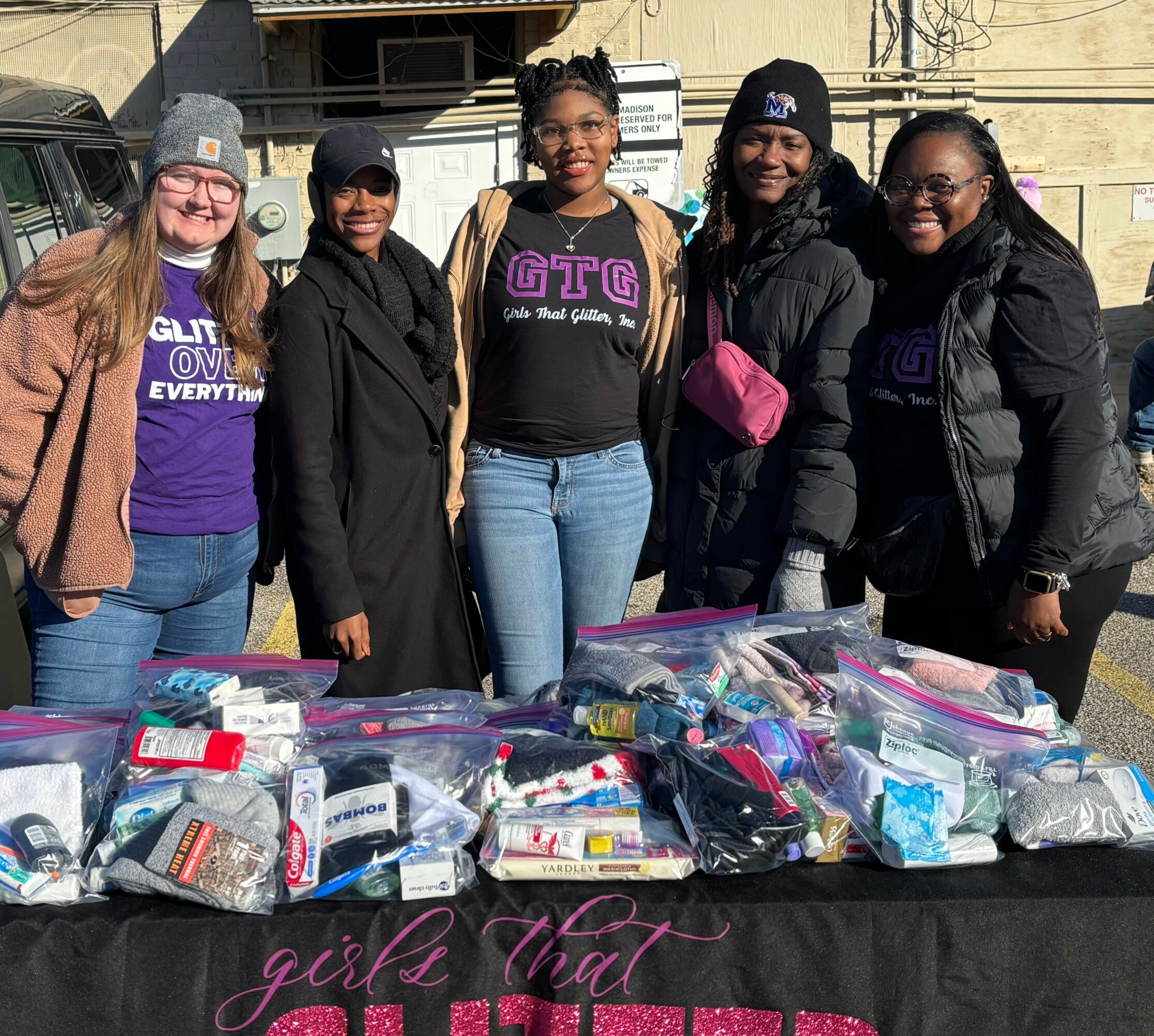 Five women stand behind a table with bags of supplies outdoors, smiling, wearing casual clothing and beanies.