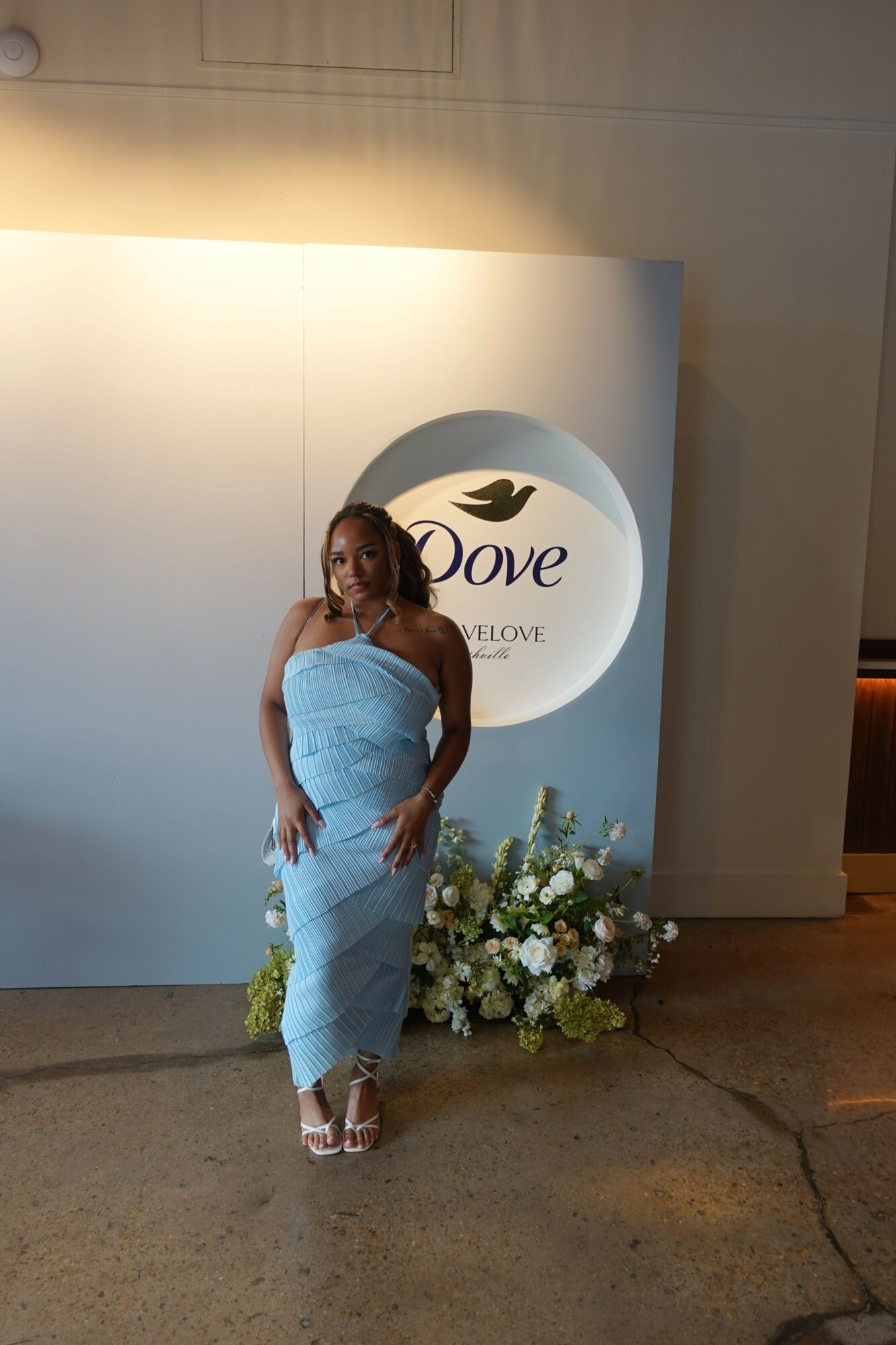 Woman in a light blue dress standing next to a floral arrangement and a Dove sign, facing sideways.