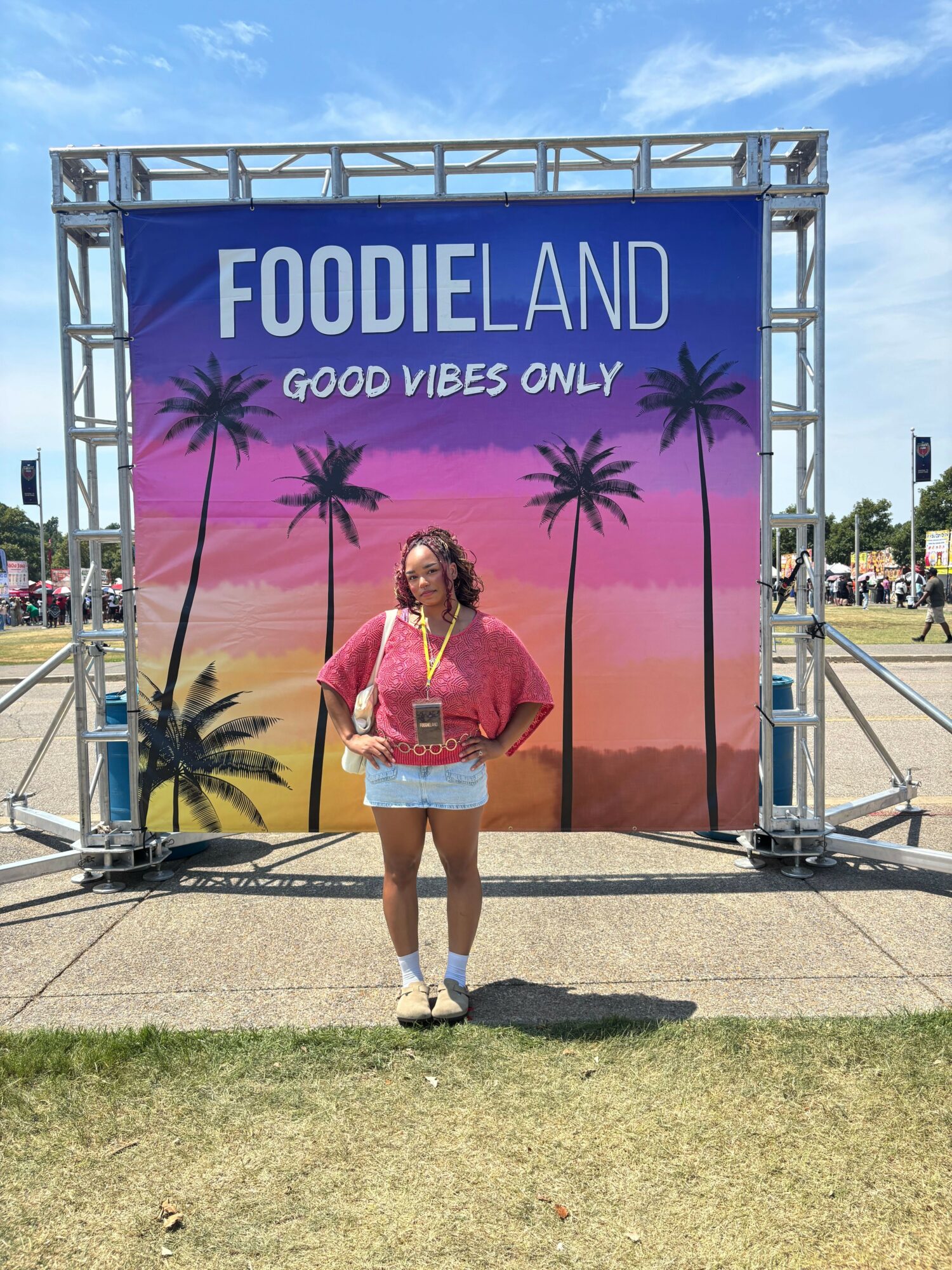 Young woman standing in front of a colorful food and vibe sign with palm trees, outdoors on a sunny day.