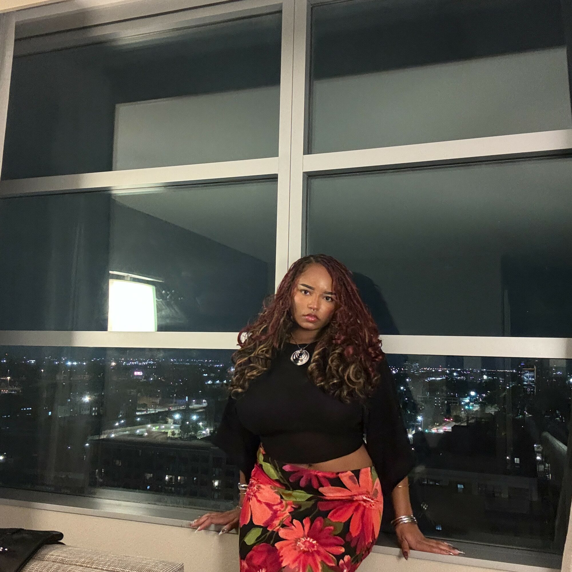 Young woman with curly hair in a black top and floral skirt standing by a large window at night, city lights visible outside.