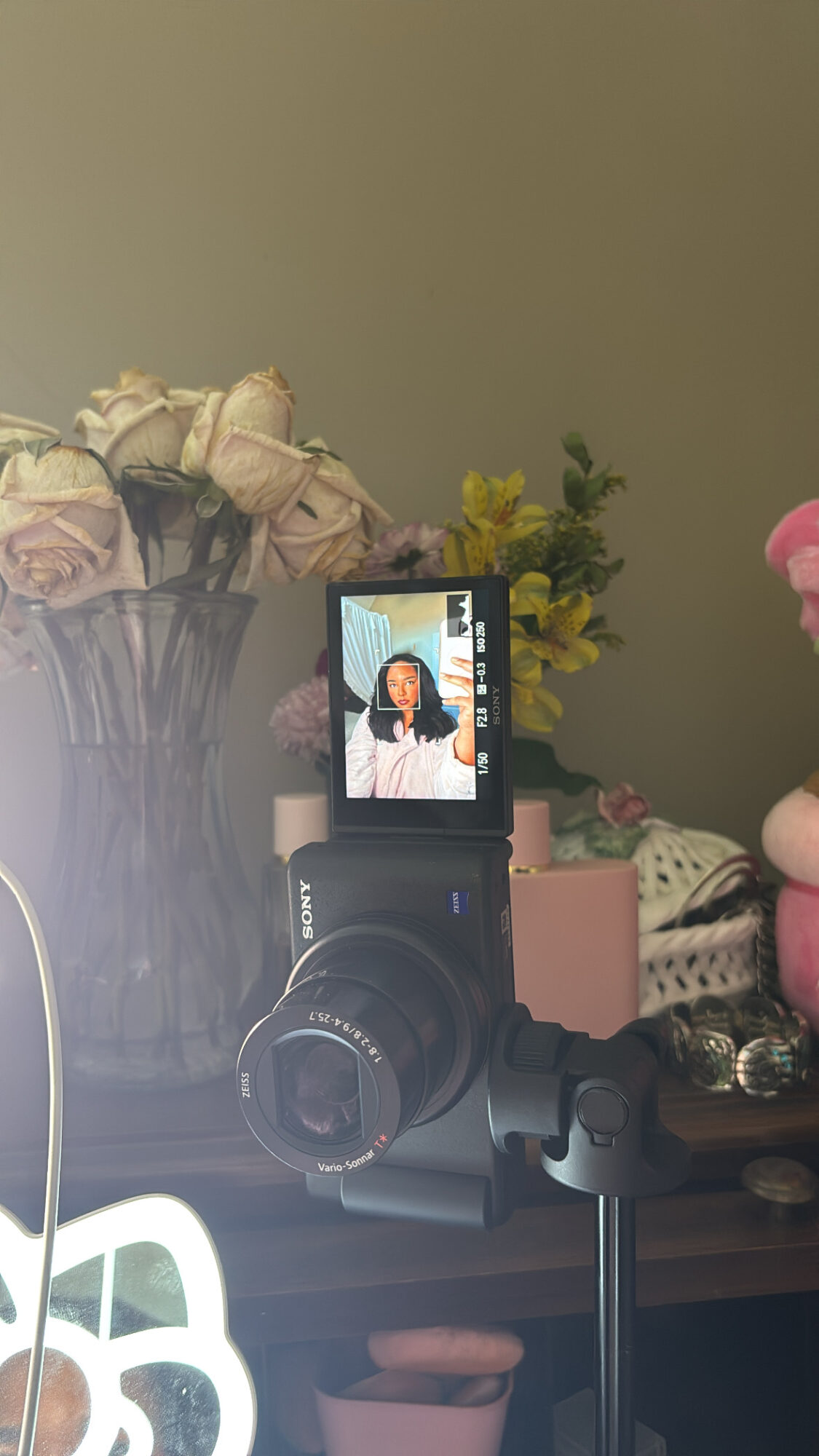 Camera on a tripod captures a woman taking a selfie, with flowers and a green wall in the background.