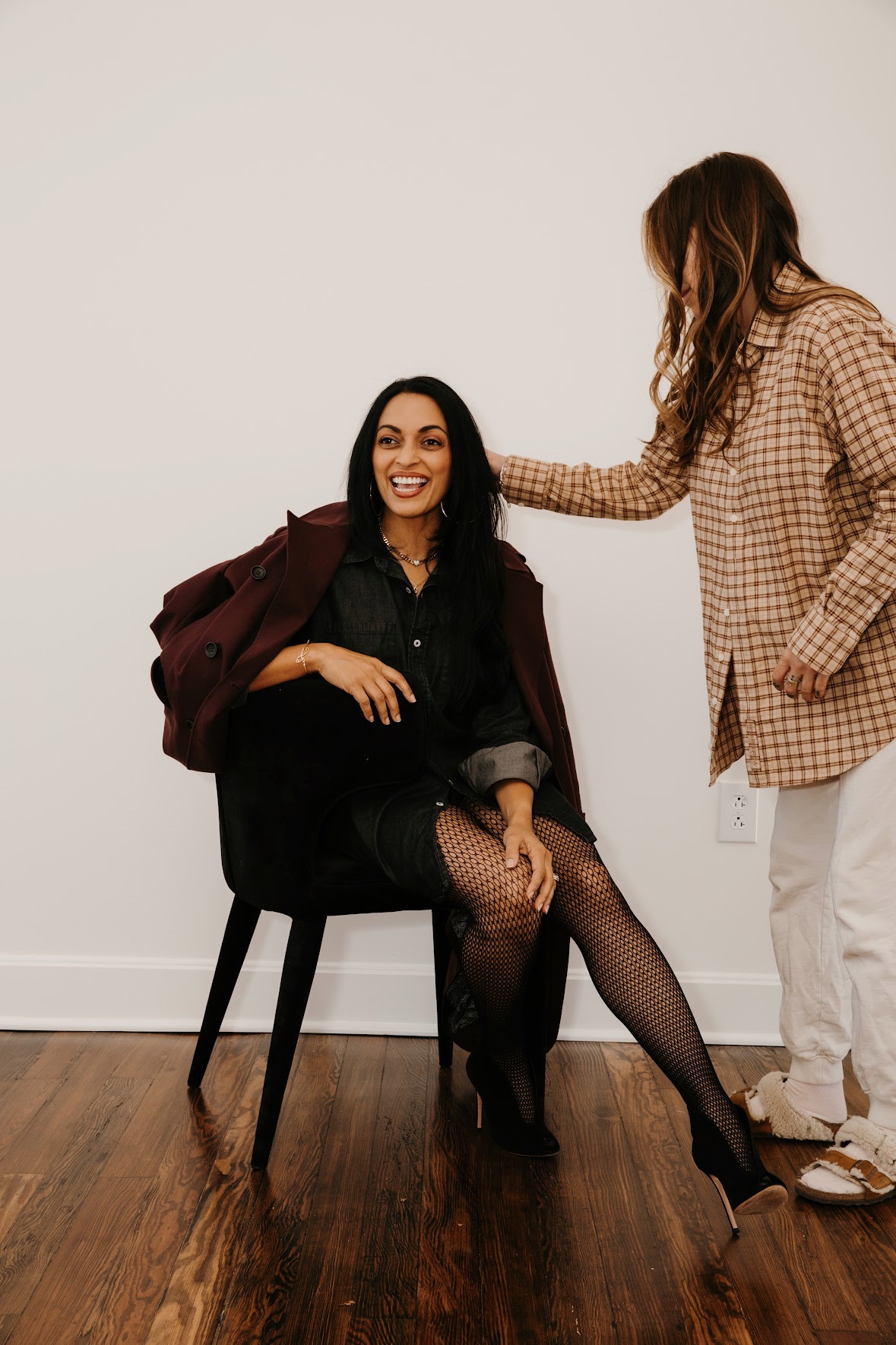 Two women in casual clothing interact indoors; one seated on a chair, smiling, the other standing and touching her shoulder.