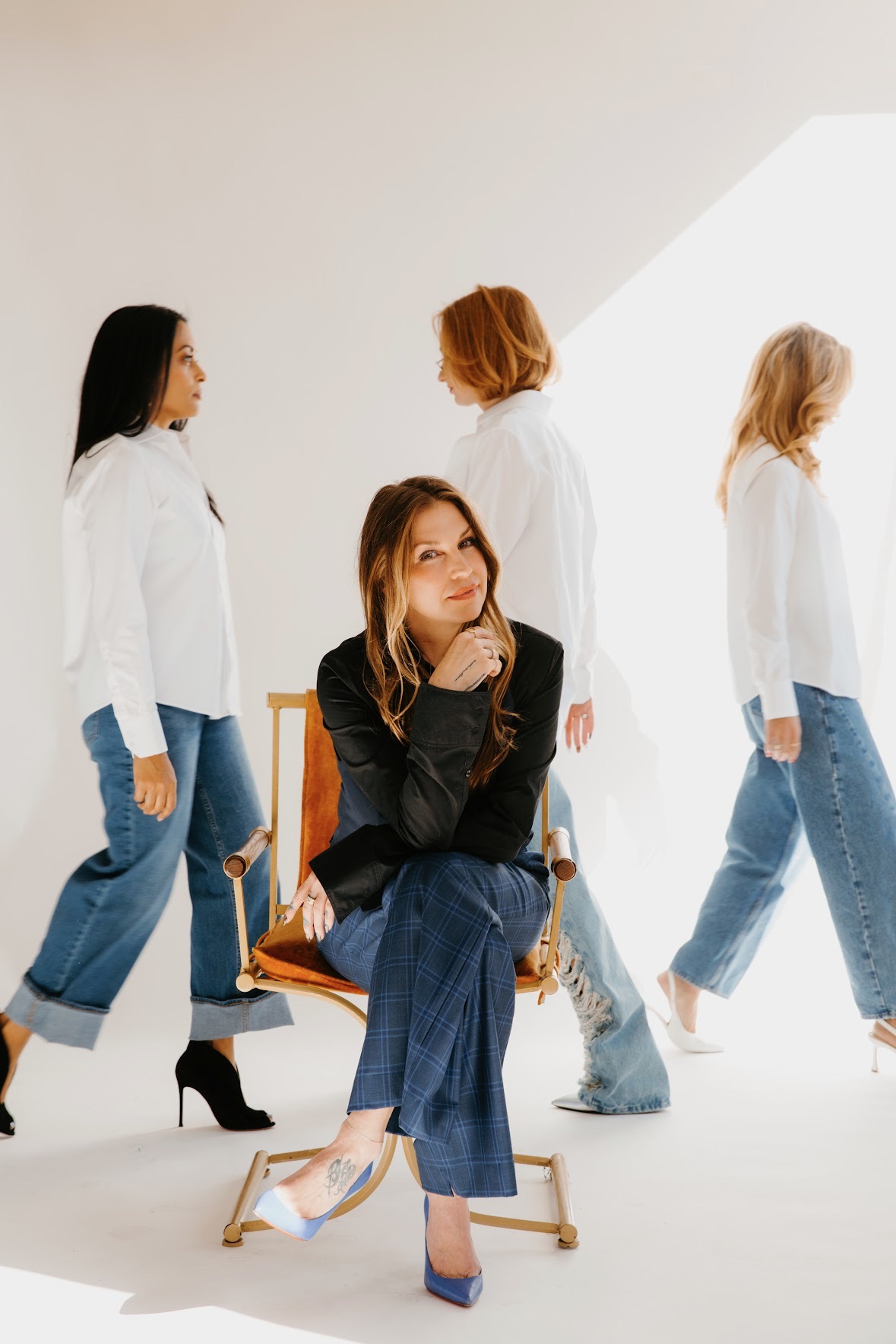 Four women in white shirts and jeans, one sitting on a chair, in a bright room with a sloped ceiling.
