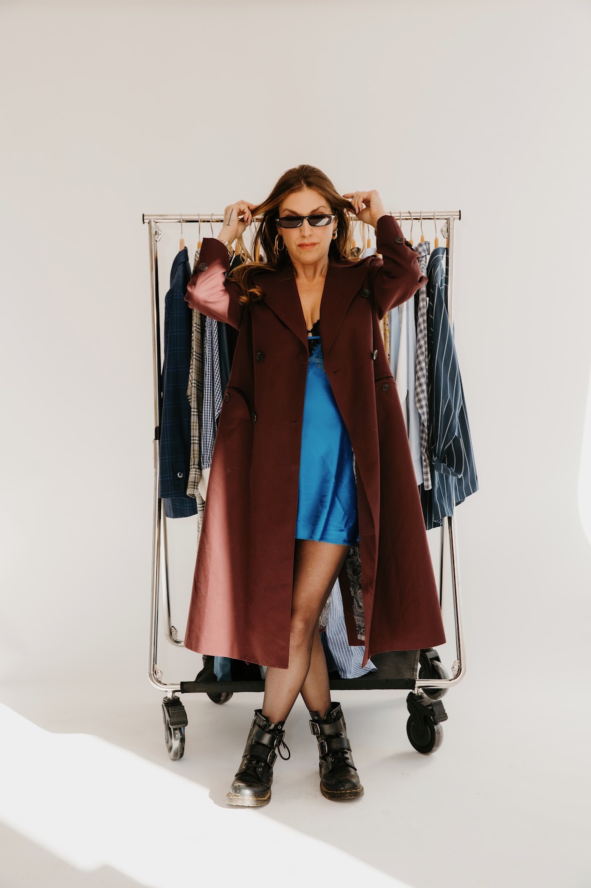 Woman with sunglasses and long coat standing in front of a clothing rack, wearing a blue dress and black boots.