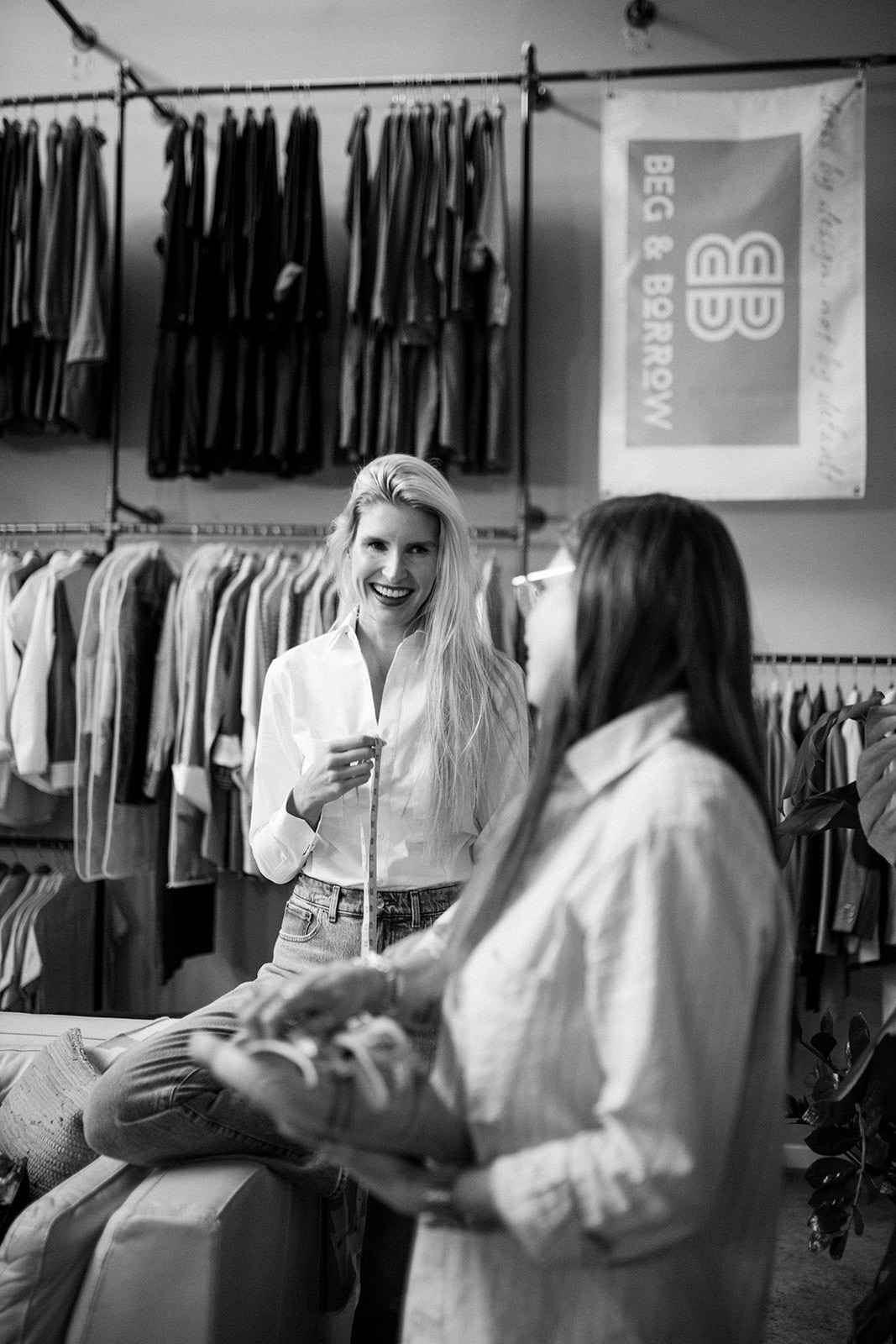 Two women in a clothing store, one smiling and holding a phone, the other holding a bag, surrounded by racks of clothes.