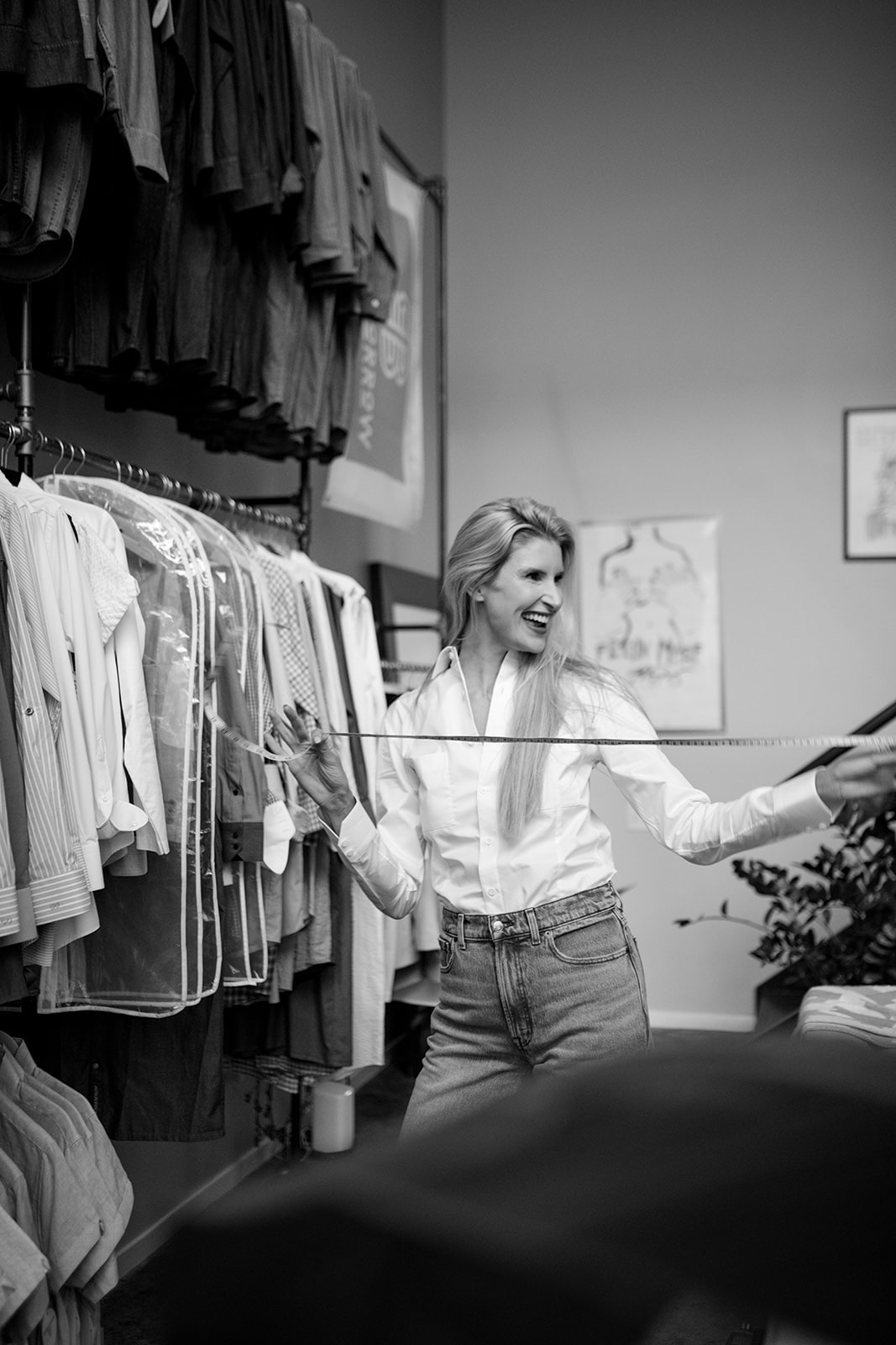 Woman shopping in clothing store, smiling, holding a hanger, surrounded by racks of clothes, in a well-lit room.