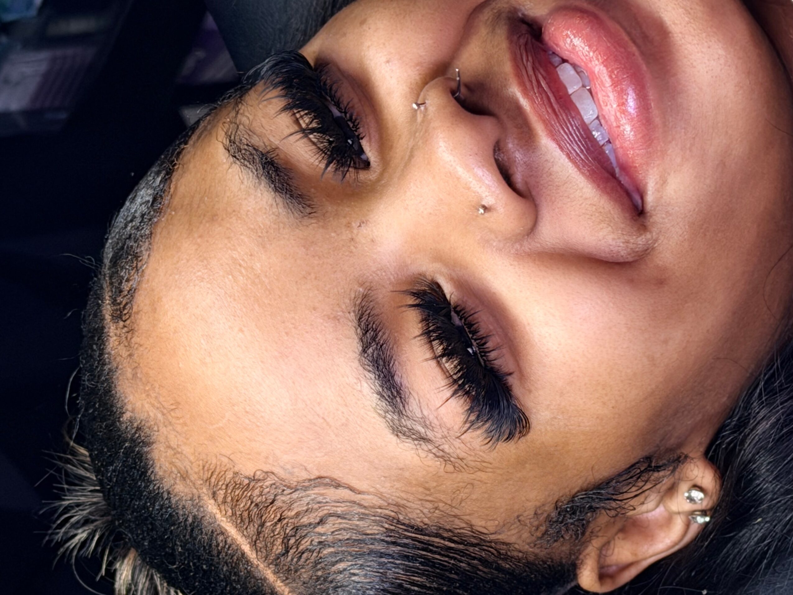 Close-up of a person with closed eyes, long eyelashes, glossy lips, and an ear stud.