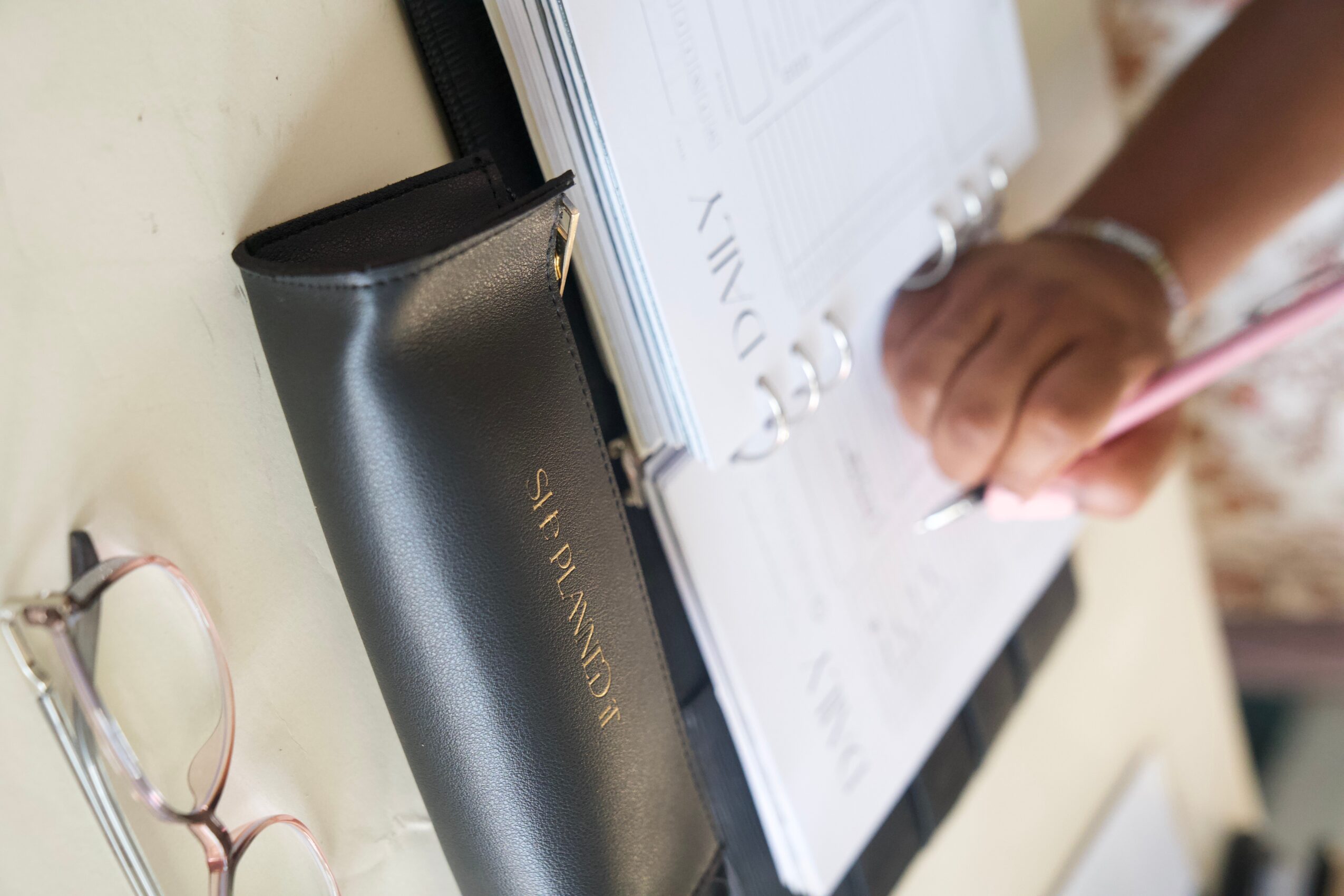 A hand writes in an open planner beside a black planner cover and eyeglasses.